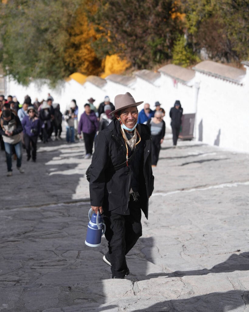 Ein lächelnder Mann mit Fedora und schwarzer Jacke trägt eine blaue Thermoskanne eine Steintreppe hinauf.