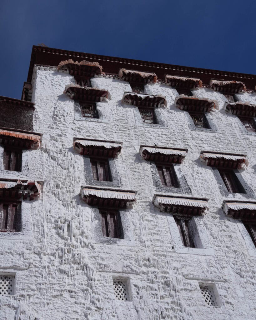 Weißes mehrstöckiges Gebäude mit dunklen Fenstern und überhängenden Dachvorsprüngen vor blauem Himmel.