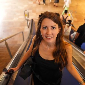 Eine Frau mit langen braunen Haaren steht auf einer Rolltreppe und lächelt in einem öffentlichen Innenraum in die Kamera – ein spontaner Moment, der perfekt für ihren Weltreise-Blog ist.