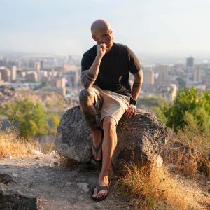 Ein Mann in Shorts und Flip-Flops sitzt an einem sonnigen Tag auf einem großen Felsen mit Blick auf die Stadt – und hält den perfekten Moment für seinen Weltreise-Blog fest.